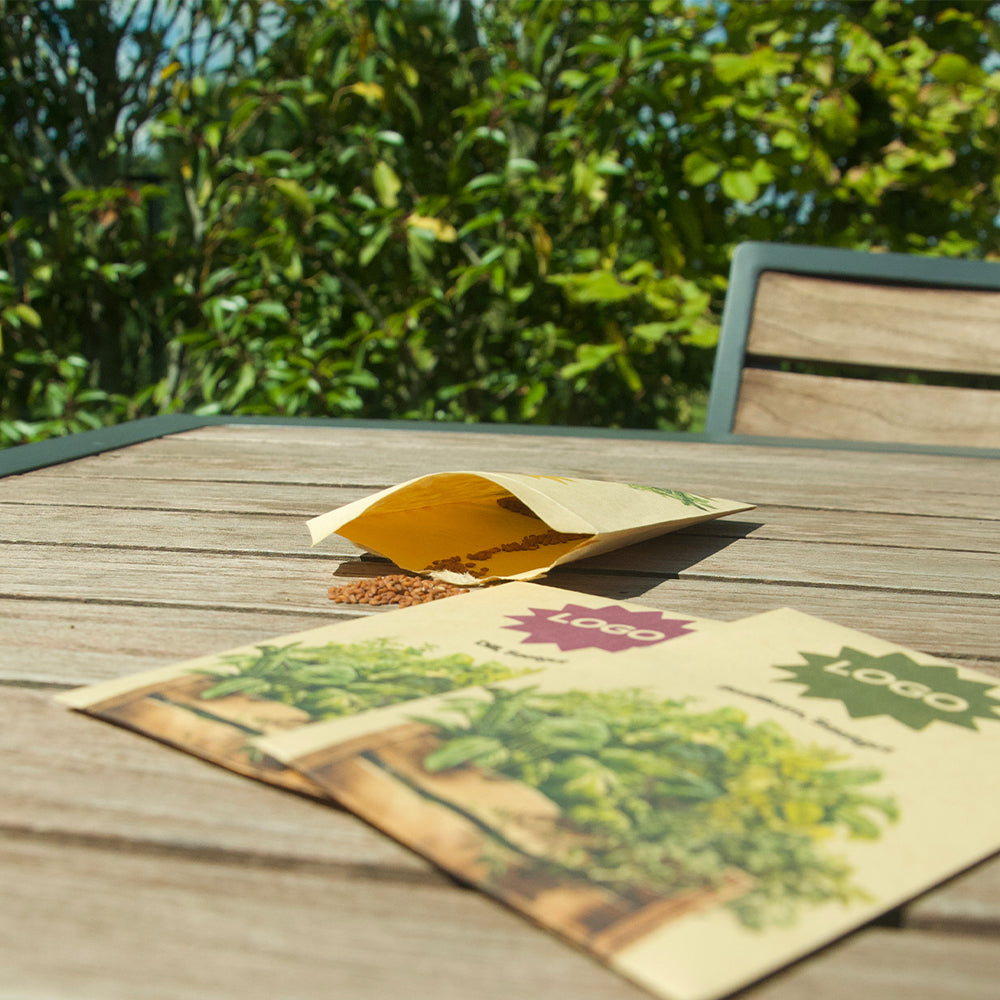 Kräutersamentütchen als Werbeartikel mit Logo auf Holztisch im Garten, zwei Verpackungen im Vordergrund, eine geöffnete Tüte mit sichtbaren Samen im Hintergrund